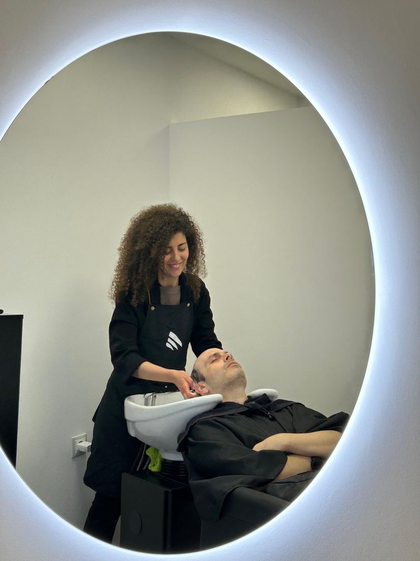 Lavado de cabello a un cliente, reflejado en el espejo redondo de la cabina.